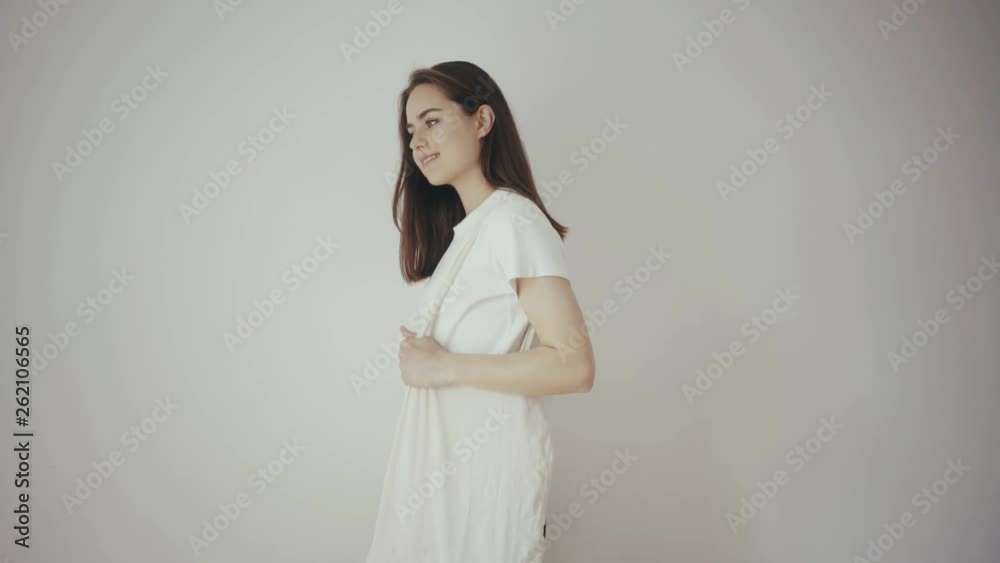 Young hipster smiling girl with white blank cotton shopper tote bag wearing white t-shirt and black jeans, mock-up of blank bag, white wall on the background, concept of shopping Young hipster smiling girl with white blank cotton shopper tote bag wearing white t-shirt and black jeans, mock-up of blank bag, white wall on the background, concept of shopping