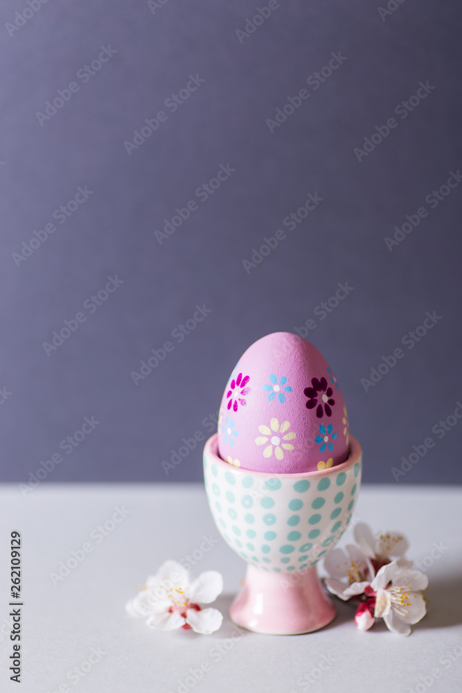 Closeup colorful painted Easter egg in vibrant modern egg stand surrounded by white flowering tree branches on gray background. Closeup colorful painted Easter egg in vibrant modern egg stand surrounded by white flowering tree branches on gray background.
