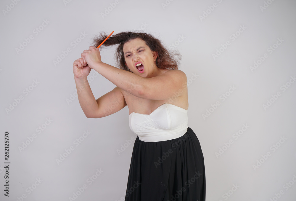 Naklejka premium emotional unhappy woman with disheveled hair tries to comb them and suffers. curvy girl stands on white background in Studio alone.
