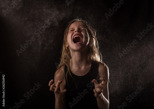 young girl with live emotions dancing under water drops. child screaming on a dark background with water splash