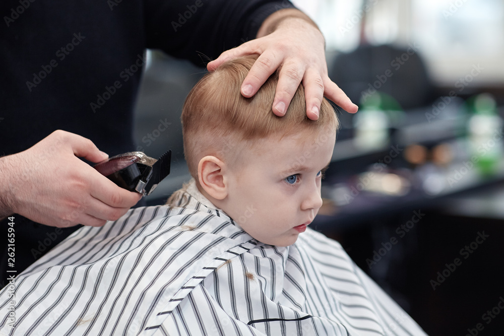 Serious cute blond baby boy with blue eyes in a barber shop having haircut  by hairdresser. Hands of stylist with tool (trimmer). Children fashion.  Indoors, dark background, copy space. Stock Photo |