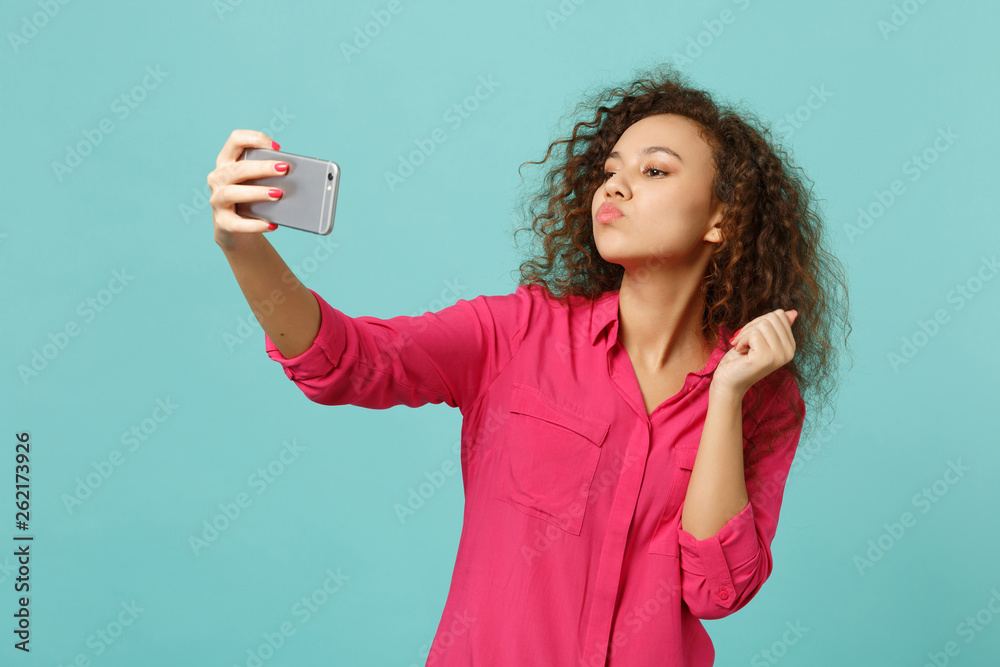 Cute african girl in casual clothes blowing sending air kiss, doing selfie shot on mobile phone isolated on blue turquoise background. People sincere emotions, lifestyle concept. Mock up copy space. Cute african girl in casual clothes blowing sending air kiss, doing selfie shot on mobile phone isolated on blue turquoise background. People sincere emotions, lifestyle concept. Mock up copy space.