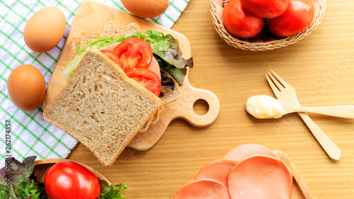 Homemade sandwich breakfast preparing.Whole wheat bread is stacked on a wooden cutting board.Fresh tomatoes, ham, eggs, spoon and fork on wooden table.Cooking ingredient.Food background concept. photo