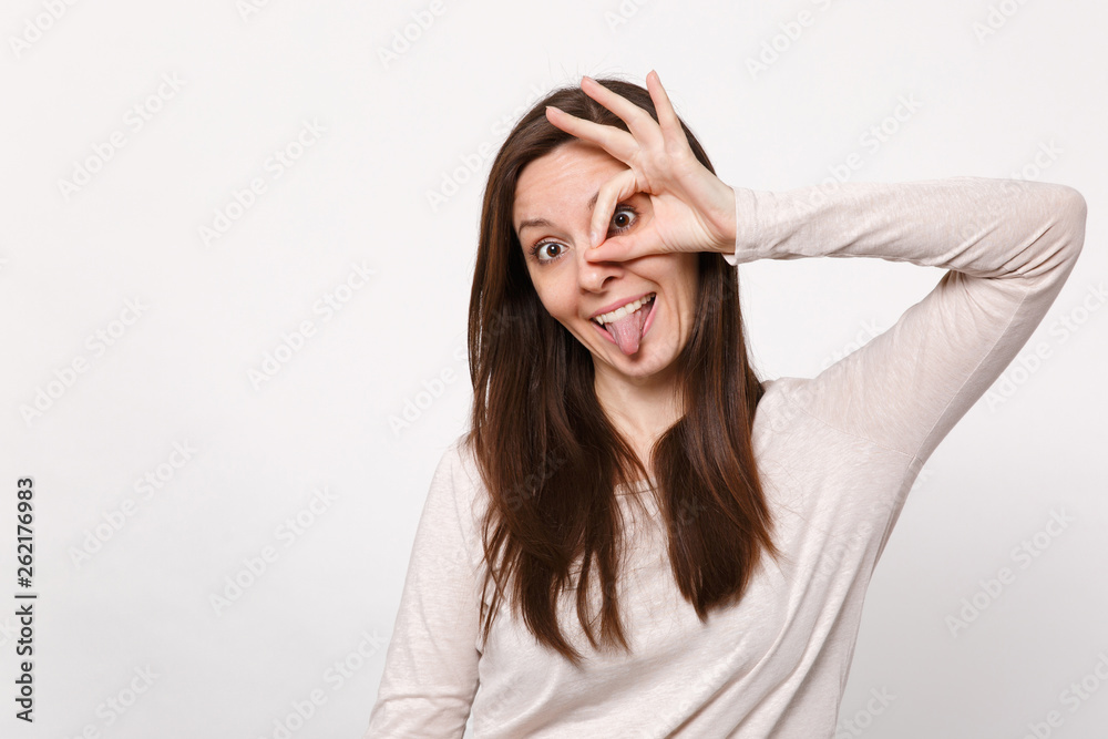 Crazy young woman in light clothes holding hands near eyes, imitating glasses or binoculars showing tongue isolated on white background. People sincere emotions, lifestyle concept. Mock up copy space. Crazy young woman in light clothes holding hands near eyes, imitating glasses or binoculars showing tongue isolated on white background. People sincere emotions, lifestyle concept. Mock up copy space.