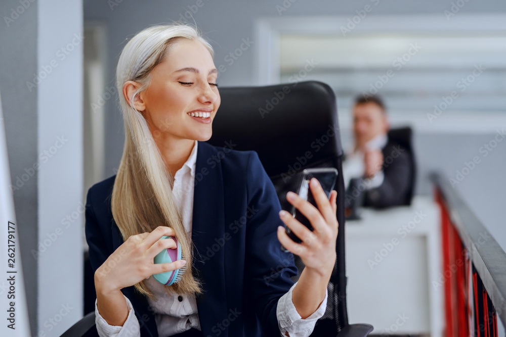 Beautiful smiling blonde Caucasian businesswoman in formal wear combing her hair while sitting in office.