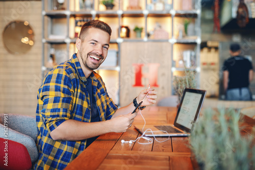 Smiling Caucasian handsome unshaven freelancer in plaid shirt putting earphones in smart phone while sitting in cafeteria. On desk laptop.