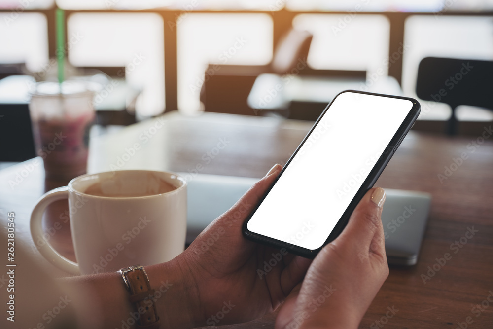 Mockup image of hands holding black mobile phone with blank desktop screen with laptop and coffee cup on the table Mockup image of hands holding black mobile phone with blank desktop screen with laptop and coffee cup on the table