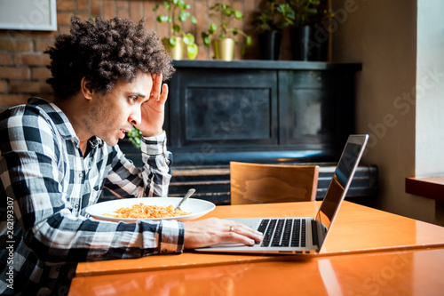 Shocked Cuban young man eating and working on laptop at restaurant photo