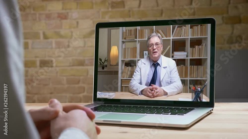 Online videocall of senior doctor in white lab coat and glasses talking seriously to his patient behind the screen. photo