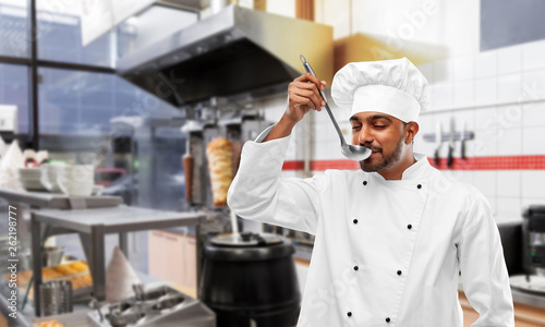 cooking, profession and people concept - happy male indian chef in toque tasting food from ladle over kebab shop kitchen background