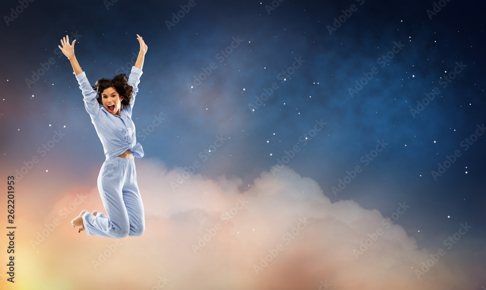 fun, people and bedtime concept - happy young woman full of energy in blue pajama jumping over starry night sky background