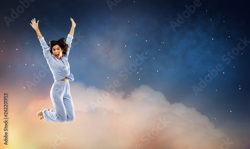 fun  people and bedtime concept - happy young woman full of energy in blue pajama jumping over starry night sky background