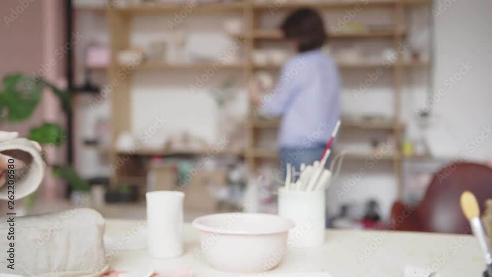 Tracking left close-up of young female ceramic artist making clay bowl in art studio, her colleague examining artworks in background Tracking left close-up of young female ceramic artist making clay bowl in art studio, her colleague examining artworks in background