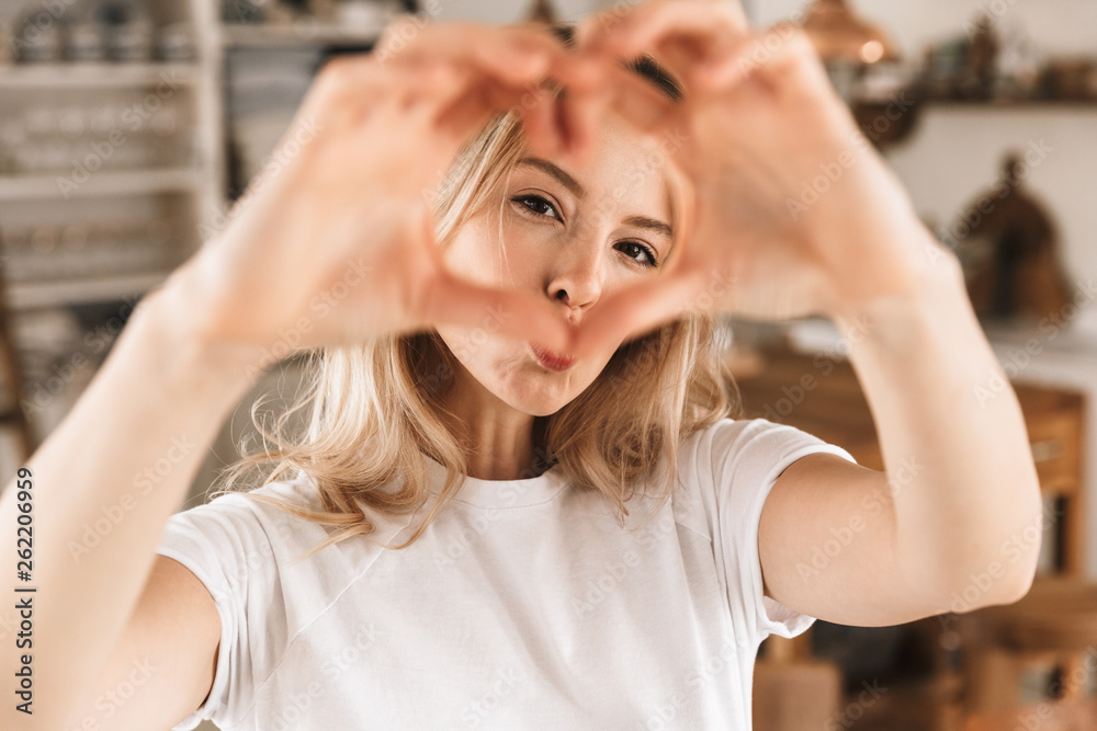 Portrait of adorable blond girl 20s wearing casual t-shirt smiling and showing heart shape with fingers while standing in stylish wooden kitchen at home Portrait of adorable blond girl 20s wearing casual t-shirt smiling and showing heart shape with fingers while standing in stylish wooden kitchen at home