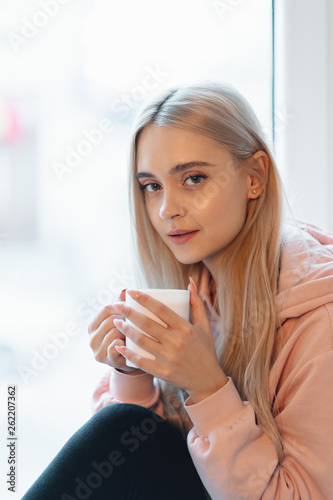 An elf alike, young girl enjoying a cup of coffee or milk while sitting by the window