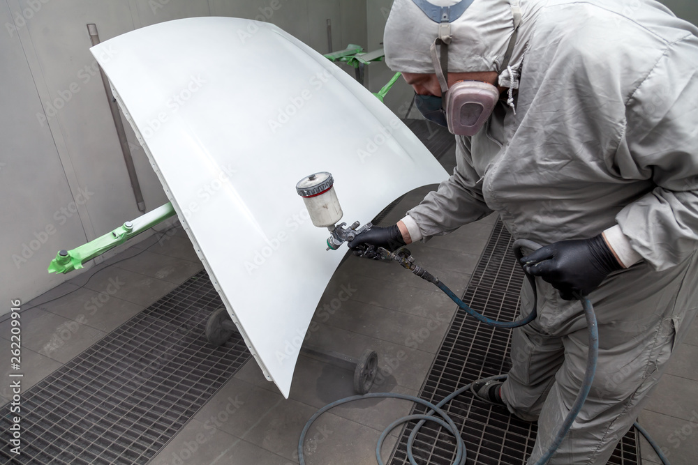 A male master worker paints with a spray gun a part of the car body in silver after being damaged at an accident. Hood from the vehicle during the repair in the workshop auto service. A male master worker paints with a spray gun a part of the car body in silver after being damaged at an accident. Hood from the vehicle during the repair in the workshop auto service.