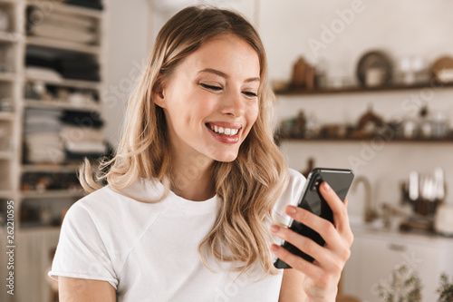 Portrait of european blond woman using smartphone while standing in stylish wooden kitchen at home