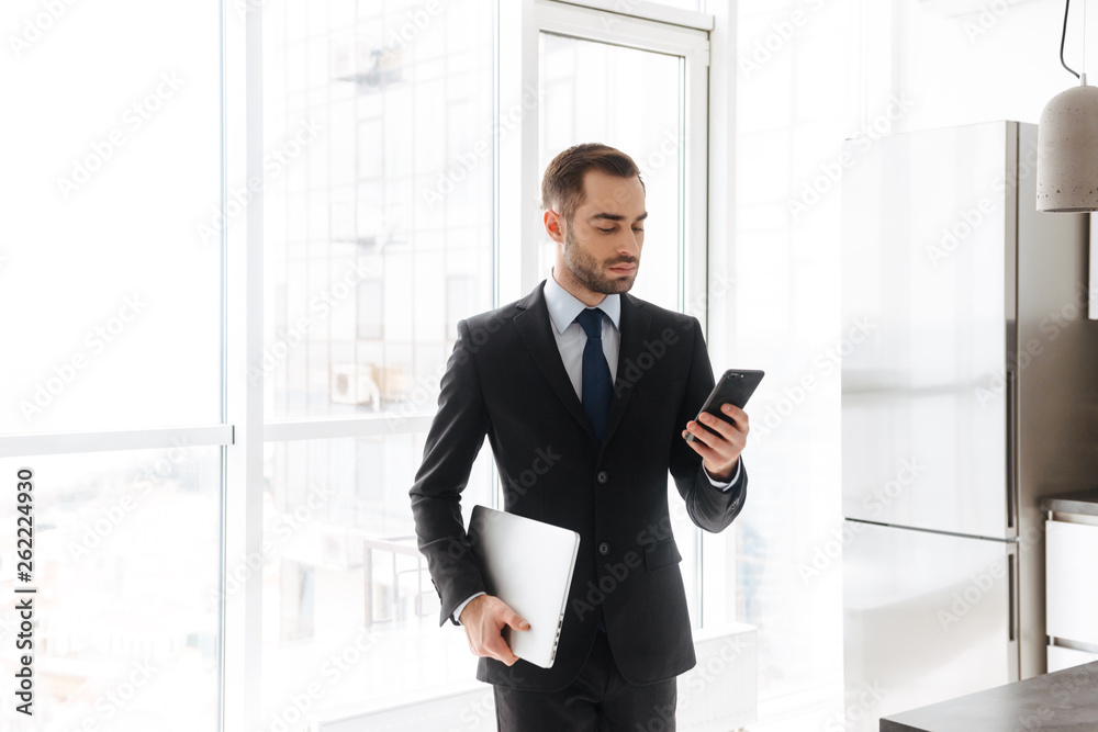 Image of handsome businessman using mobile phone while working on laptop in bright apartment Image of handsome businessman using mobile phone while working on laptop in bright apartment