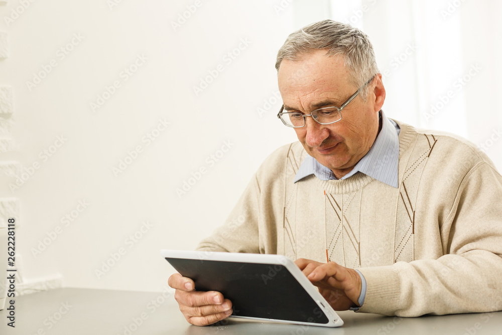 Handsome cheerful senior man using digital tablet sitting at the table in the living room. Leisure activities, spending time, happy retirement and senior lifestyle concept