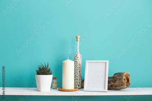 Modern room decoration with Picture frame mockup. White shelf against pastel turquoise wall with Candle and rocks in bottle. Potted succulent plant