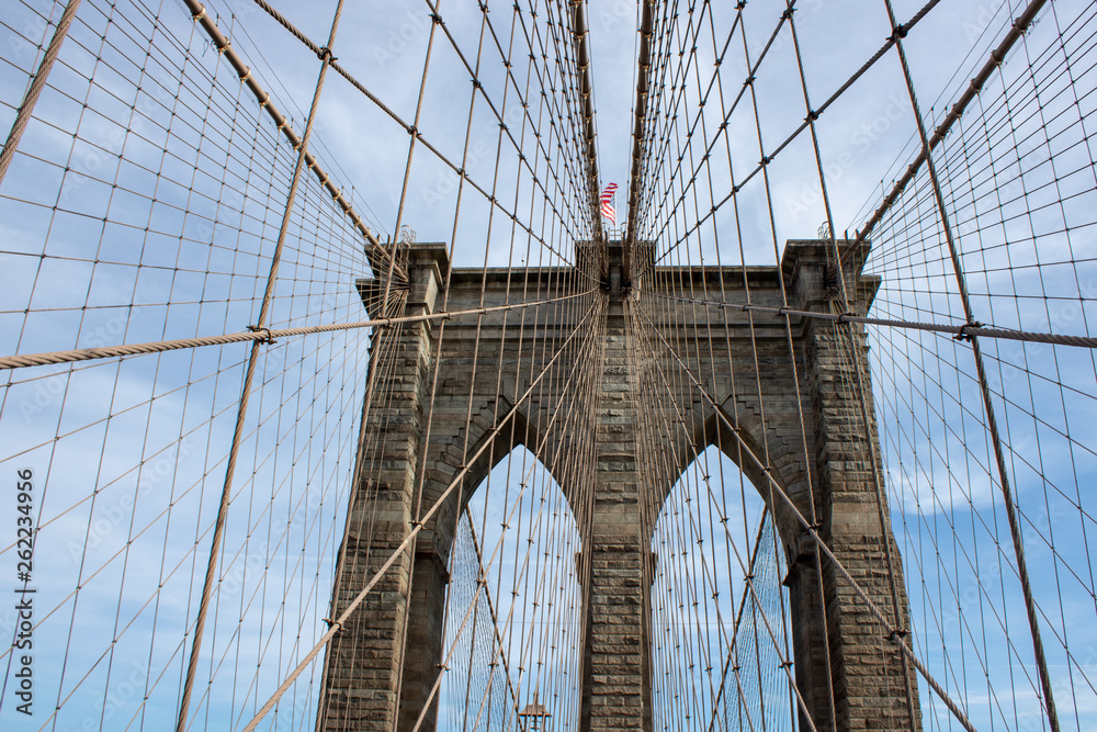 Naklejka premium The famous Brooklyn Bridge Bridge located in New York City in the United States of America showing the suspension wired and the USA flag at the top of the column on a part cloudy day with blue Skys.