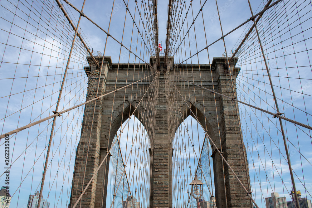 Fototapeta premium The famous Brooklyn Bridge Bridge located in New York City in the United States of America showing the suspension wired and the USA flag at the top of the column on a part cloudy day with blue Skys.