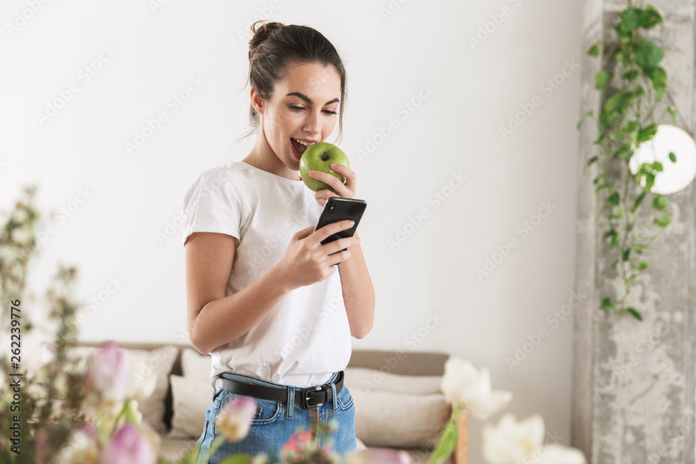 Happy beautiful young student girl eat apple posing indoors using mobile phone. Happy beautiful young student girl eat apple posing indoors using mobile phone.