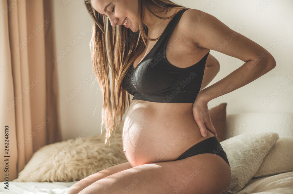 Fototapeta premium Attractive pregnant woman is sitting in bed and holding her belly. Last months of pregnancy
