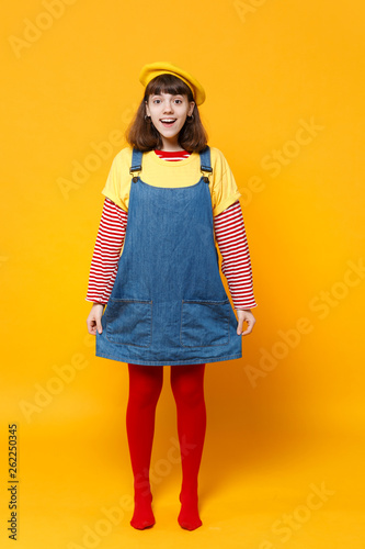 Full length portrait of excited girl teenager in french beret and denim sundress standing isolated on yellow wall background in studio. People sincere emotions, lifestyle concept. Mock up copy space.