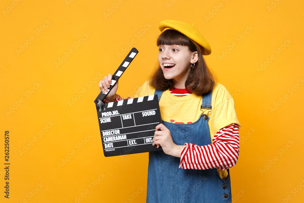 Smiling girl teenager in french beret looking aside, holding classic black film making clapperboard isolated on yellow wall background. People sincere emotions, lifestyle concept. Mock up copy space.