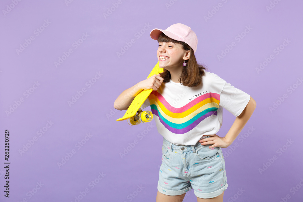 Portrait of smiling teen girl in vivid clothes holding yellow skateboard, looking aside isolated on violet pastel background in studio. People sincere emotions, lifestyle concept. Mock up copy space. Portrait of smiling teen girl in vivid clothes holding yellow skateboard, looking aside isolated on violet pastel background in studio. People sincere emotions, lifestyle concept. Mock up copy space.