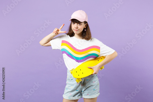 Portrait of pretty teen girl in vivid clothes with yellow skateboard showing victory sign isolated on violet pastel background in studio. People sincere emotions lifestyle concept. Mock up copy space.