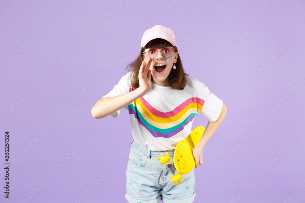 Excited teen girl in vivid clothes, heart eyeglasses hold skateboard, screaming with hand gesture isolated on violet pastel background. People sincere emotions, lifestyle concept. Mock up copy space. Excited teen girl in vivid clothes, heart eyeglasses hold skateboard, screaming with hand gesture isolated on violet pastel background. People sincere emotions, lifestyle concept. Mock up copy space.