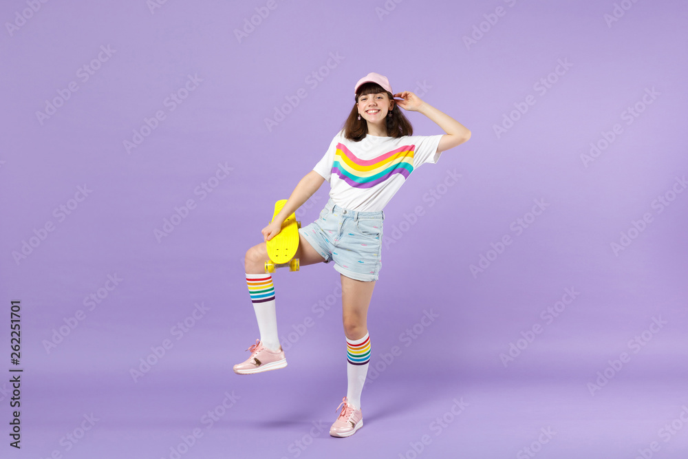 Portrait of funny teen girl in vivid clothes standing, holding yellow skateboard isolated on violet pastel wall background in studio. People sincere emotions, lifestyle concept. Mock up copy space.