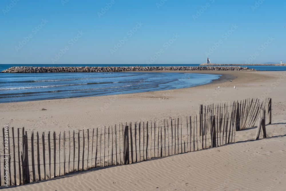 Sandy beach at La Grande-Motte city with a wooden fence at the foreground and a stone jetty on the background. France 2019.