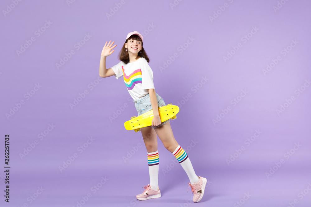 Side view of pleasant teen girl hold skateboard looking back waving greeting with hand as notices someone isolated on violet background. People sincere emotions, lifestyle concept. Mock up copy space. Side view of pleasant teen girl hold skateboard looking back waving greeting with hand as notices someone isolated on violet background. People sincere emotions, lifestyle concept. Mock up copy space.