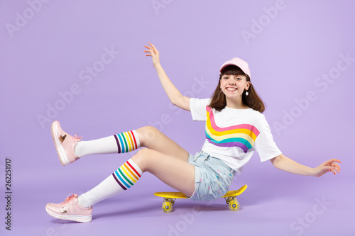 Funny cute teen girl in vivid clothes sitting on yellow skateboard, spreading hands isolated on violet pastel wall background in studio. People sincere emotions lifestyle concept. Mock up copy space.