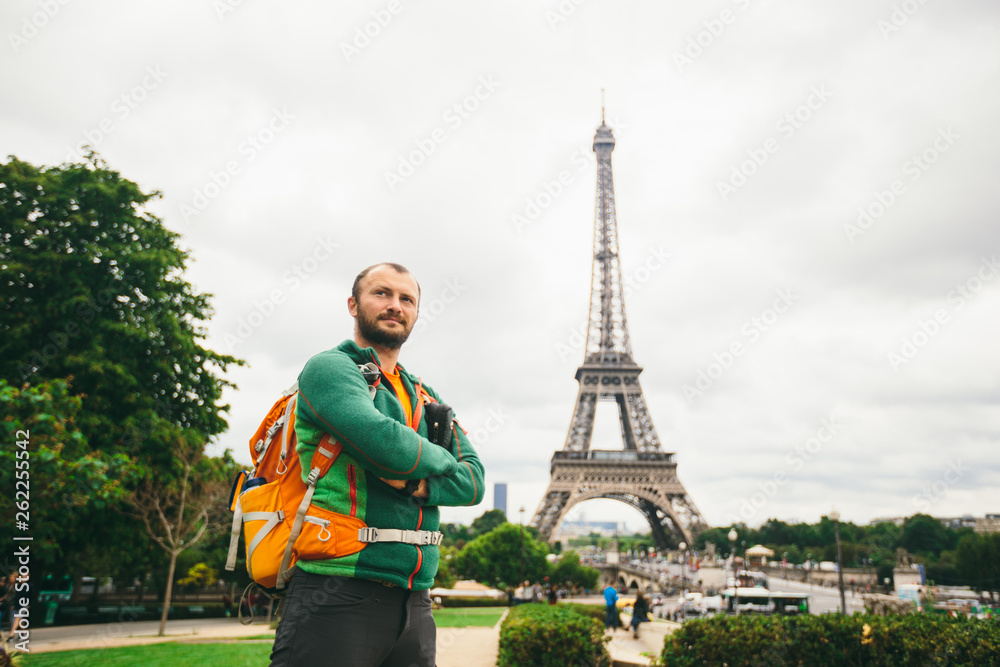 A young Caucasian Caucasian man with an orange backpack and a photo camera in his hands is standing in France, paris against the background of the Eiffel Tower