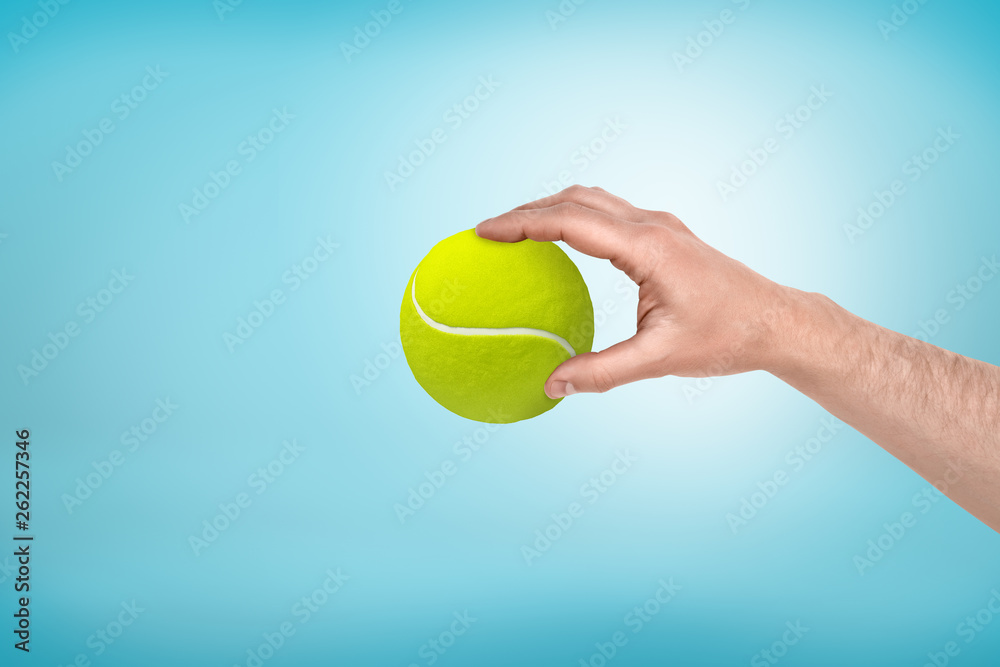 Male hand holding small tennis ball between fingers on blue background