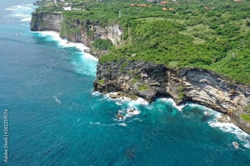 Aerial view of green cliff and blue ocean with breaking waves in Uluwatu Temple, Bali, Indonesia.