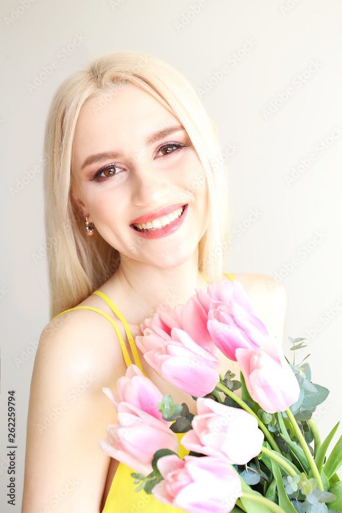 Studio portrait of gorgeous young blonde woman with long straight hair wearing bright yellow dress and holding bouquet of many tender pink tulips. Gray isolated background, copy space, close up. Studio portrait of gorgeous young blonde woman with long straight hair wearing bright yellow dress and holding bouquet of many tender pink tulips. Gray isolated background, copy space, close up.