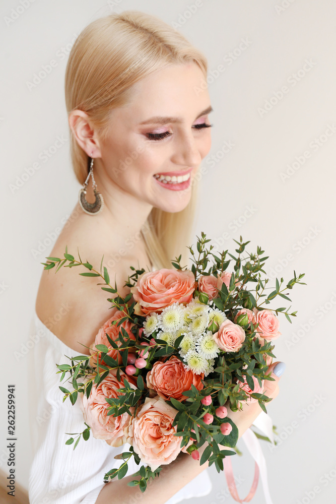 Studio portrait of gorgeous young blonde woman with long straight hair wearing soft beige dress and holding bouquet of many tender pale pink tulips. Gray isolated background, copy space, close up.