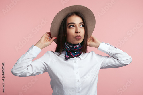 Fashionable young girl wearing white shirt and stylish scarf, holding her hat and looking away over pink studio wall