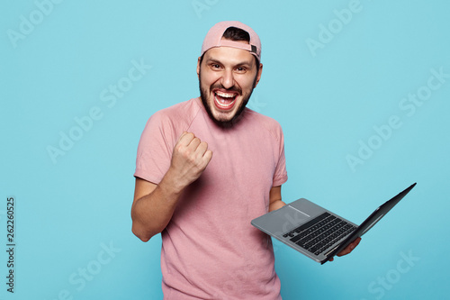 Joyful stylish student wearing pink baseball cap standing and holding laptop, fist up in success on blue background.