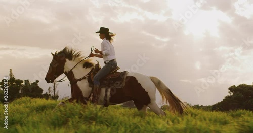 Cowgirl horseback riding at sunset in green field, brown and white horse cantering with rider photo