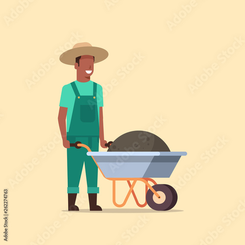 gardener man pushing wheelbarrow full of earth compost african american male farmer working in garden wearing overalls gardening concept full length