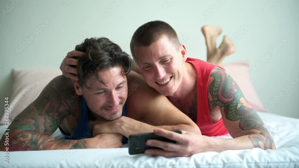 Young gay male couple in bed together using phone. Sharing good memory. Happy gay friendship, relationship, pride concept. Young gay male couple in bed together using phone. Sharing good memory. Happy gay friendship, relationship, pride concept.