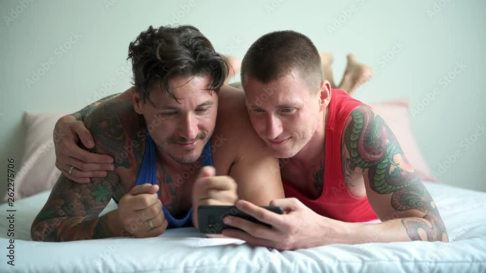 Young gay male couple in bed together using phone. Talking. Happy gay friendship, relationship, pride concept. Young gay male couple in bed together using phone. Talking. Happy gay friendship, relationship, pride concept.