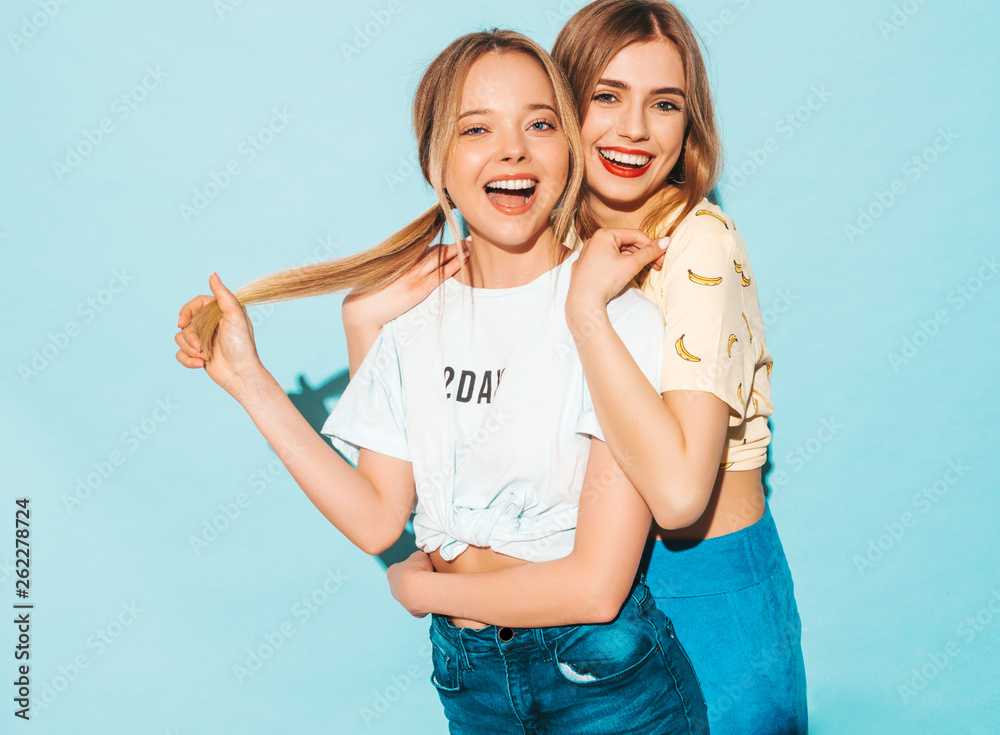 Two young beautiful smiling blond hipster girls in trendy summer colorful T-shirt clothes. Sexy carefree women posing near blue wall. Positive models having fun and hugging Two young beautiful smiling blond hipster girls in trendy summer colorful T-shirt clothes. Sexy carefree women posing near blue wall. Positive models having fun and hugging