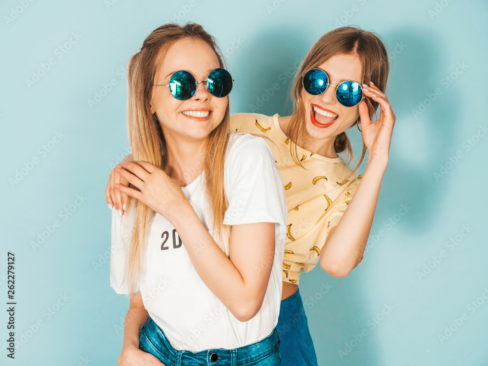 Two young beautiful smiling blond hipster girls in trendy summer colorful  clothes. Sexy carefree women posing near blue wall in round sunglasses. Positive models having fun